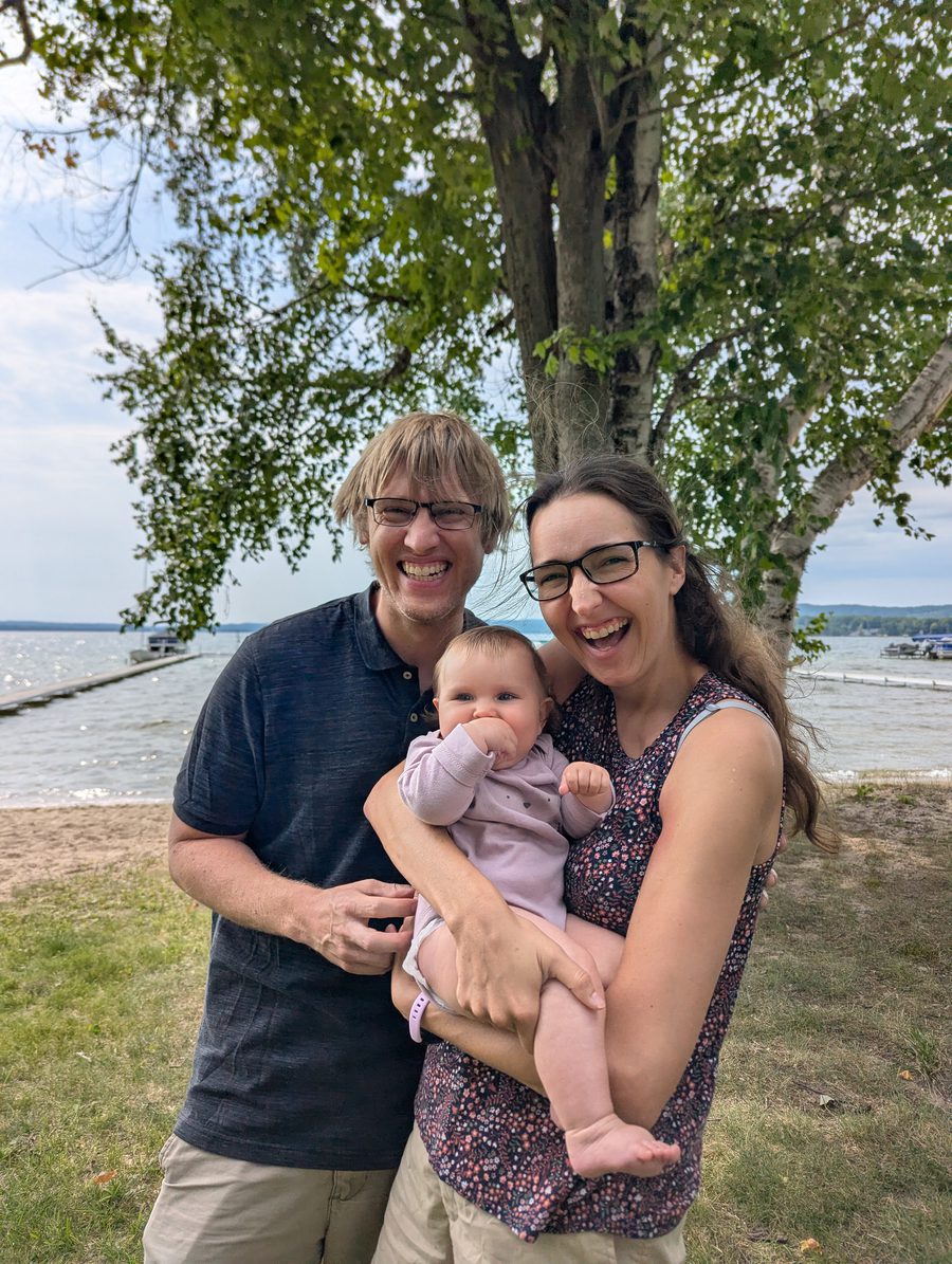 Luke and Katelyn holding their baby daughter on a grassy lakeshore