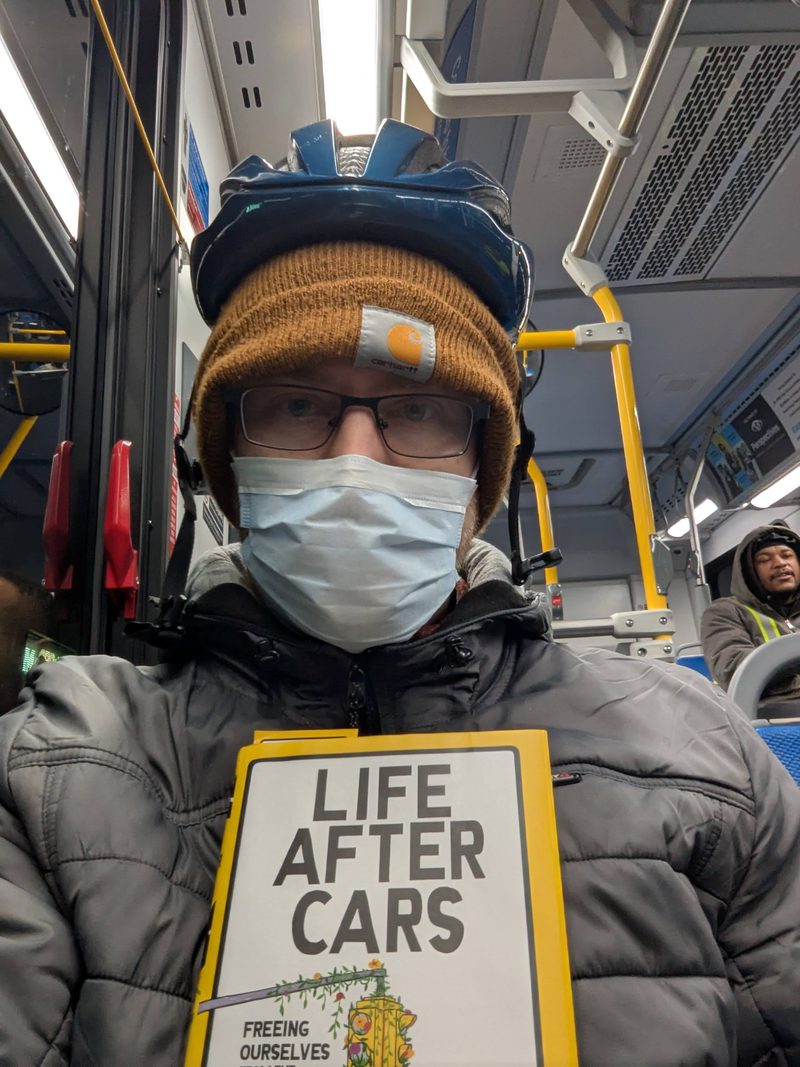 Luke in a mask and bike helmet on a bus, holding the book Life After Cars